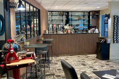 Salle de restaurant lumineuse avec tables dressées, chaises noires et vue sur une rue arborée à travers de grandes baies…