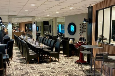 Salle de restaurant lumineuse avec tables dressées, chaises noires et vue sur une rue arborée à travers de grandes baies…