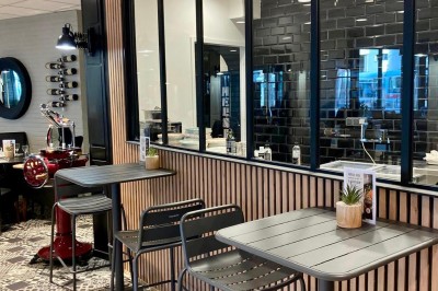 Salle de restaurant lumineuse avec tables dressées, chaises noires et vue sur une rue arborée à travers de grandes baies…
