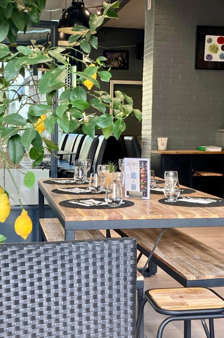 Salle de restaurant avec tables en bois dressées, chaises noires et plantes décoratives au premier plan.