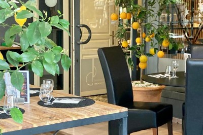 Salle de restaurant lumineuse avec tables dressées, chaises noires et vue sur une rue arborée à travers de grandes baies…