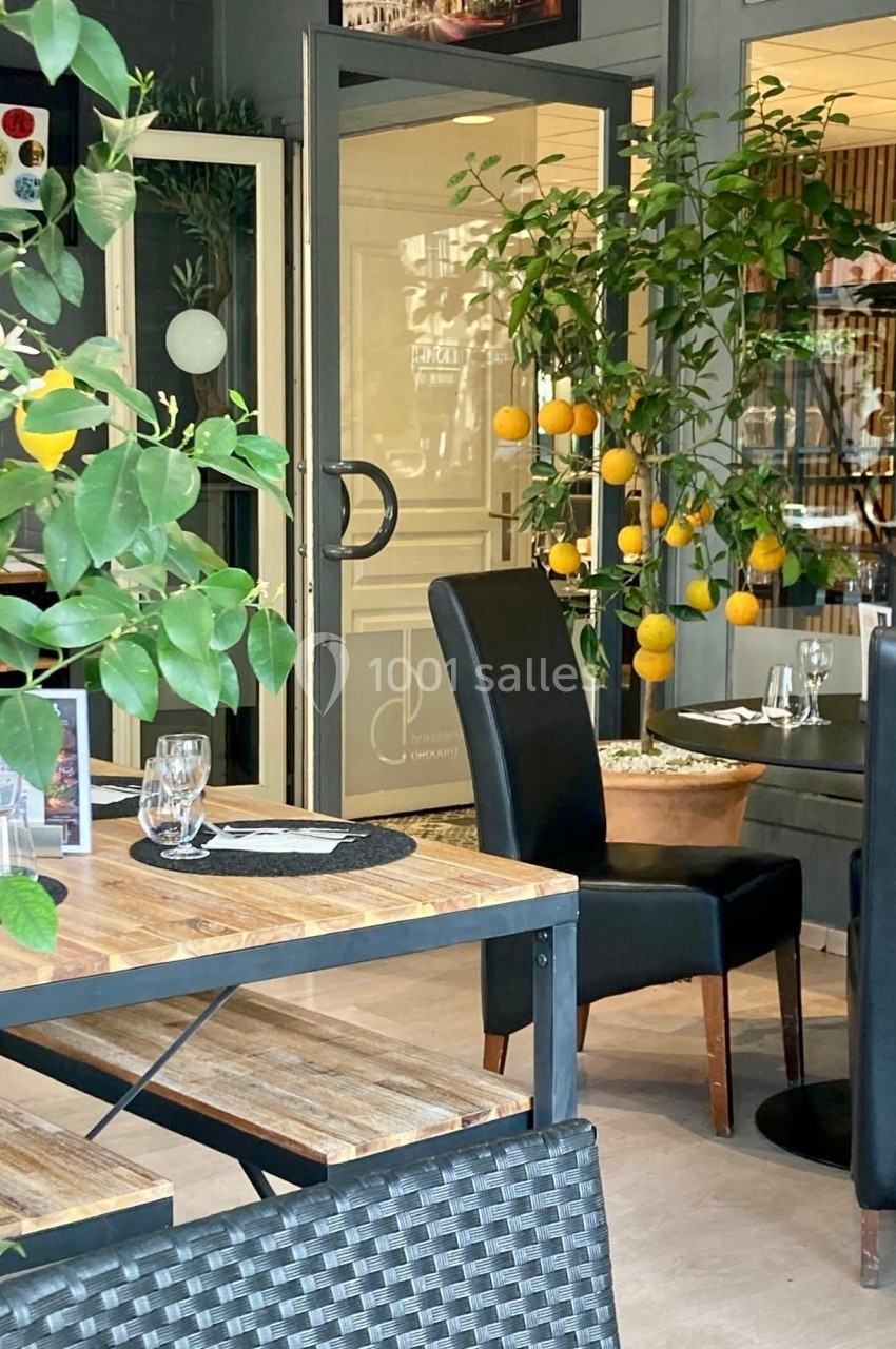 Salle de restaurant lumineuse avec tables en bois, chaises noires et citronniers en pot près des portes vitrées.