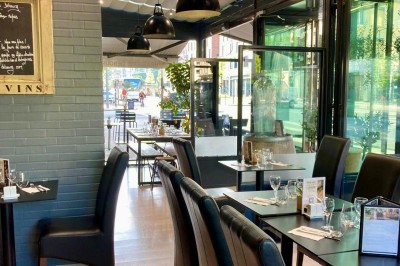 Salle de restaurant lumineuse avec tables dressées, chaises noires et vue sur une rue arborée à travers de grandes baies…