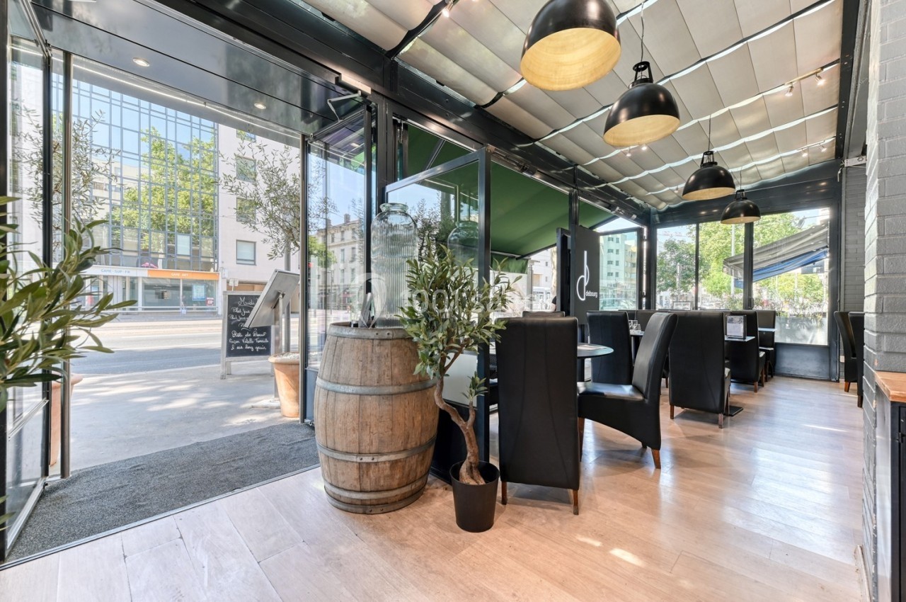 Salle de restaurant lumineuse avec baies vitrées, mobilier moderne, tonneau en bois et plantes décoratives.