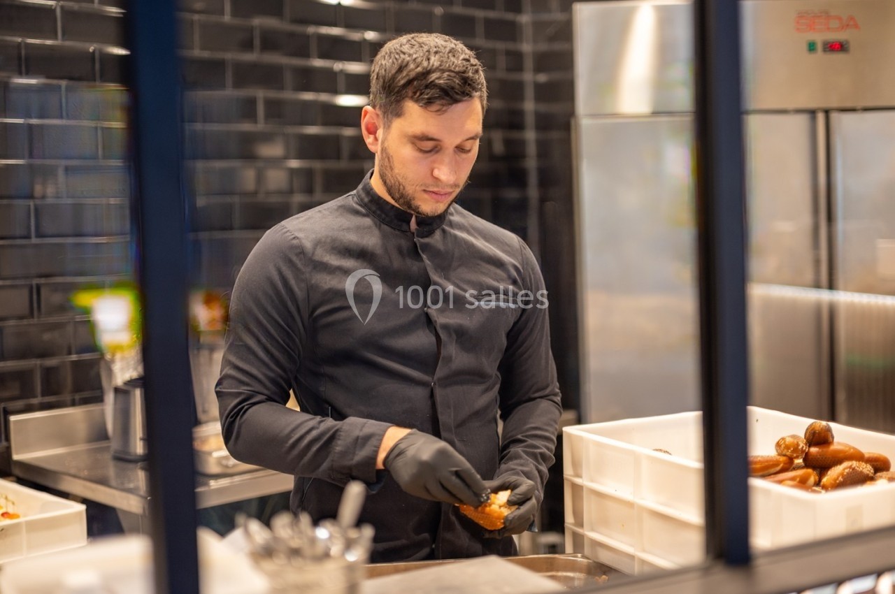 Un homme en tenue de cuisine noire prépare des aliments dans une cuisine professionnelle avec des gants noirs.