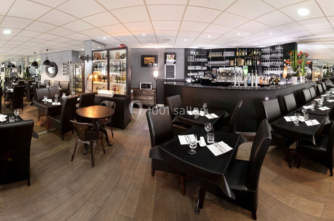 Salle de restaurant moderne avec tables dressées, chaises noires et bar en arrière-plan.