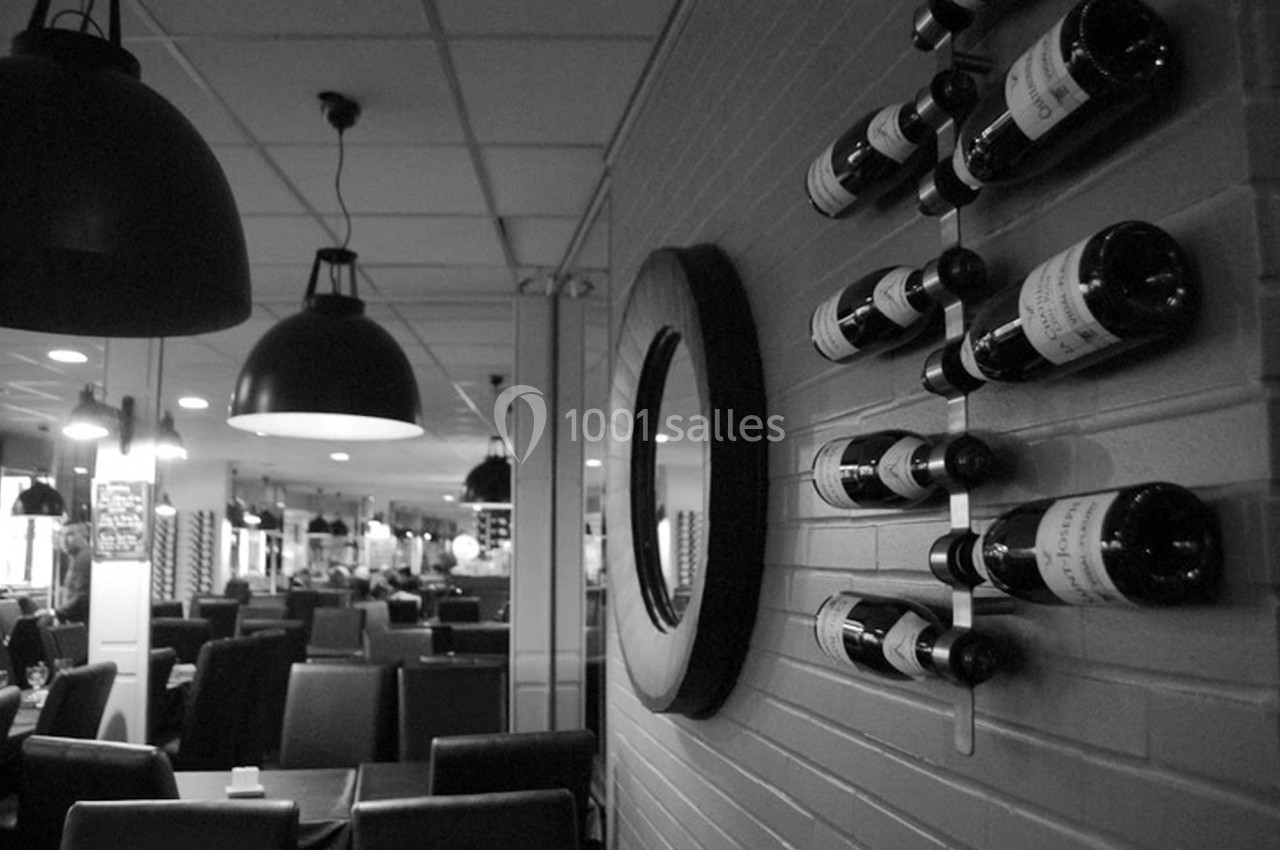Bouteilles de vin disposées sur un support mural dans un restaurant avec des tables et des luminaires suspendus.