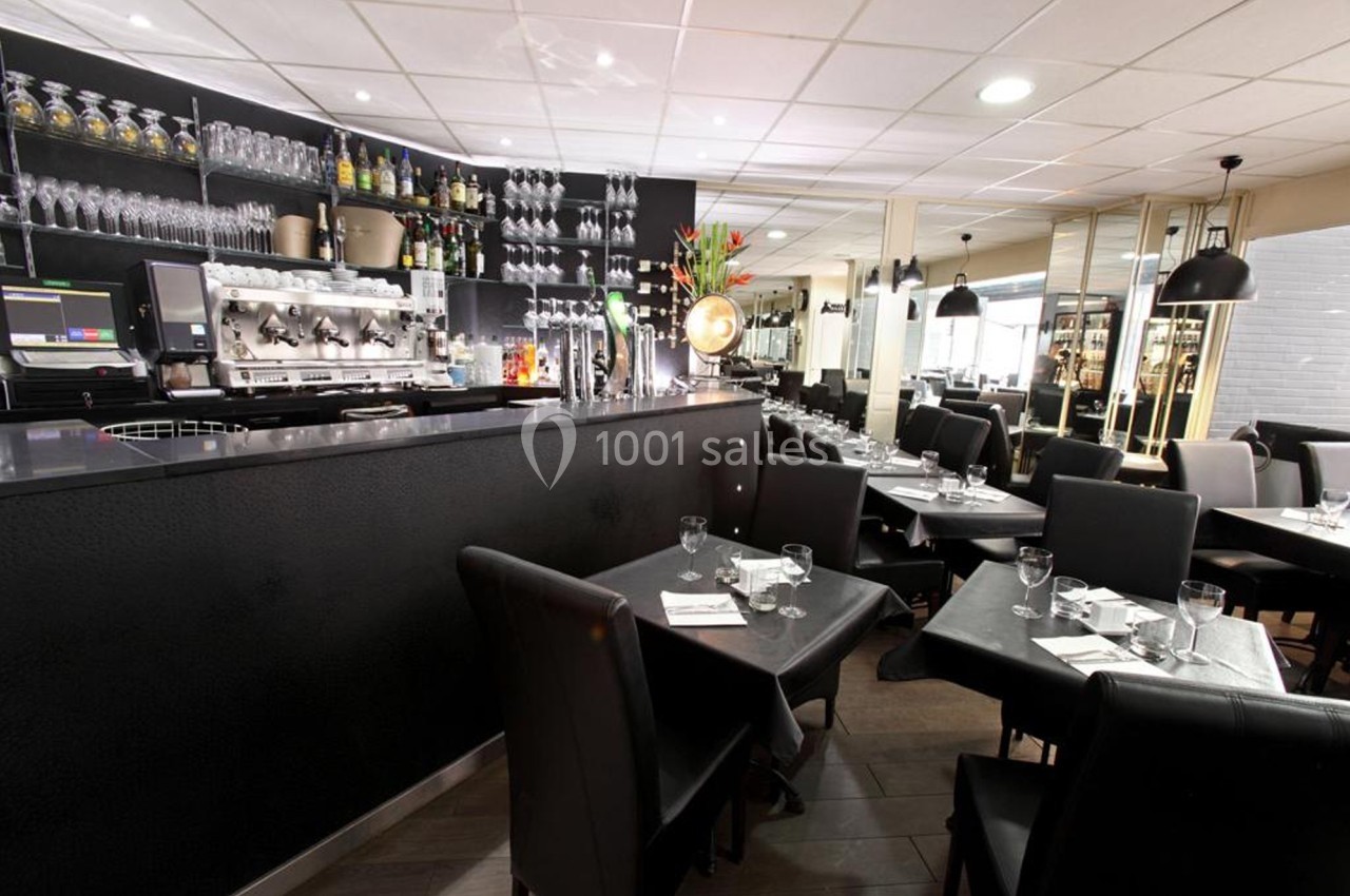 Salle de restaurant moderne avec tables dressées, chaises noires, bar équipé et éclairage chaleureux.
