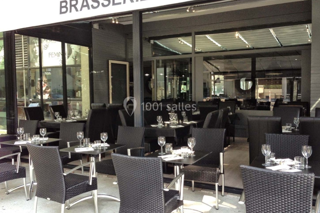 Terrasse et intérieur d'une brasserie avec des tables dressées, chaises en osier noir et grande baie vitrée.