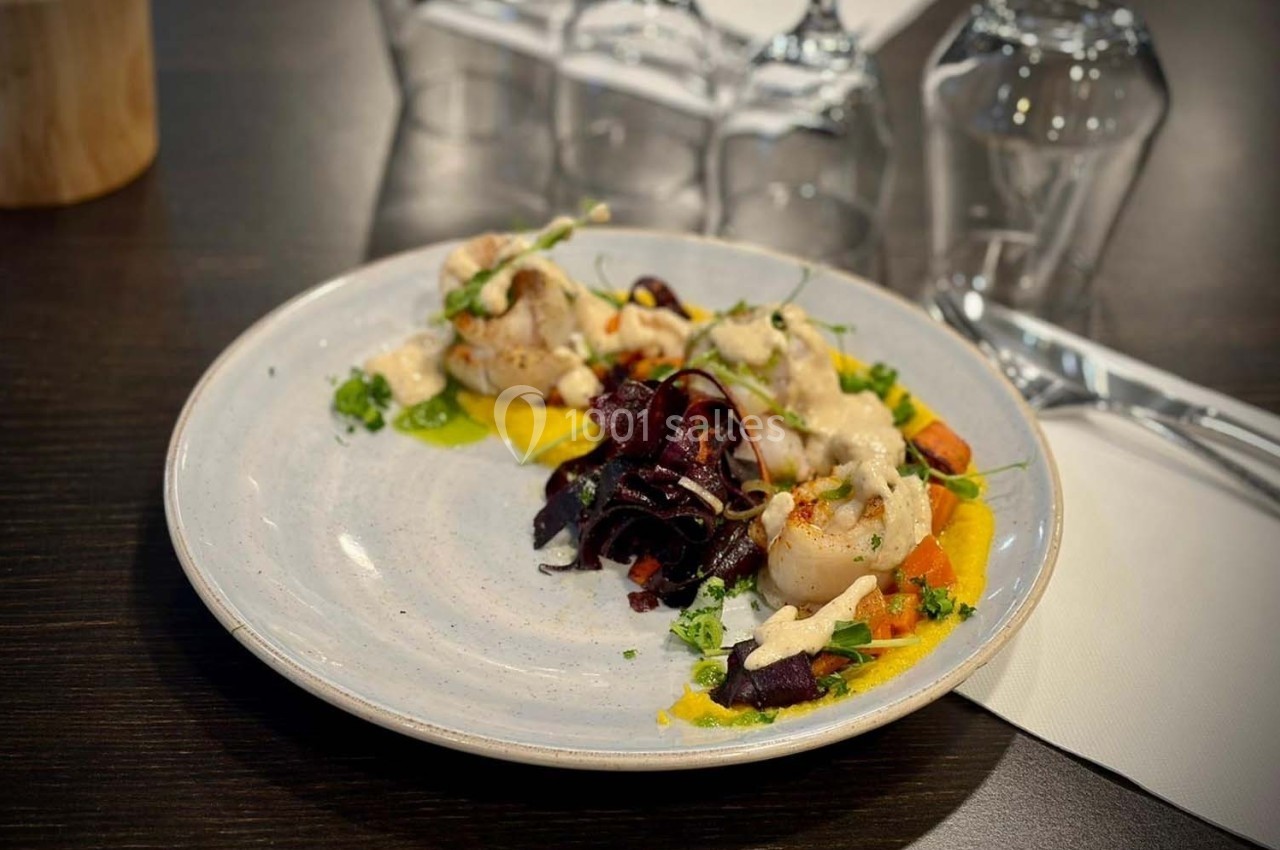 Assiette de noix de Saint-Jacques avec purée, légumes colorés et sauce, servie sur une table sombre avec couverts.