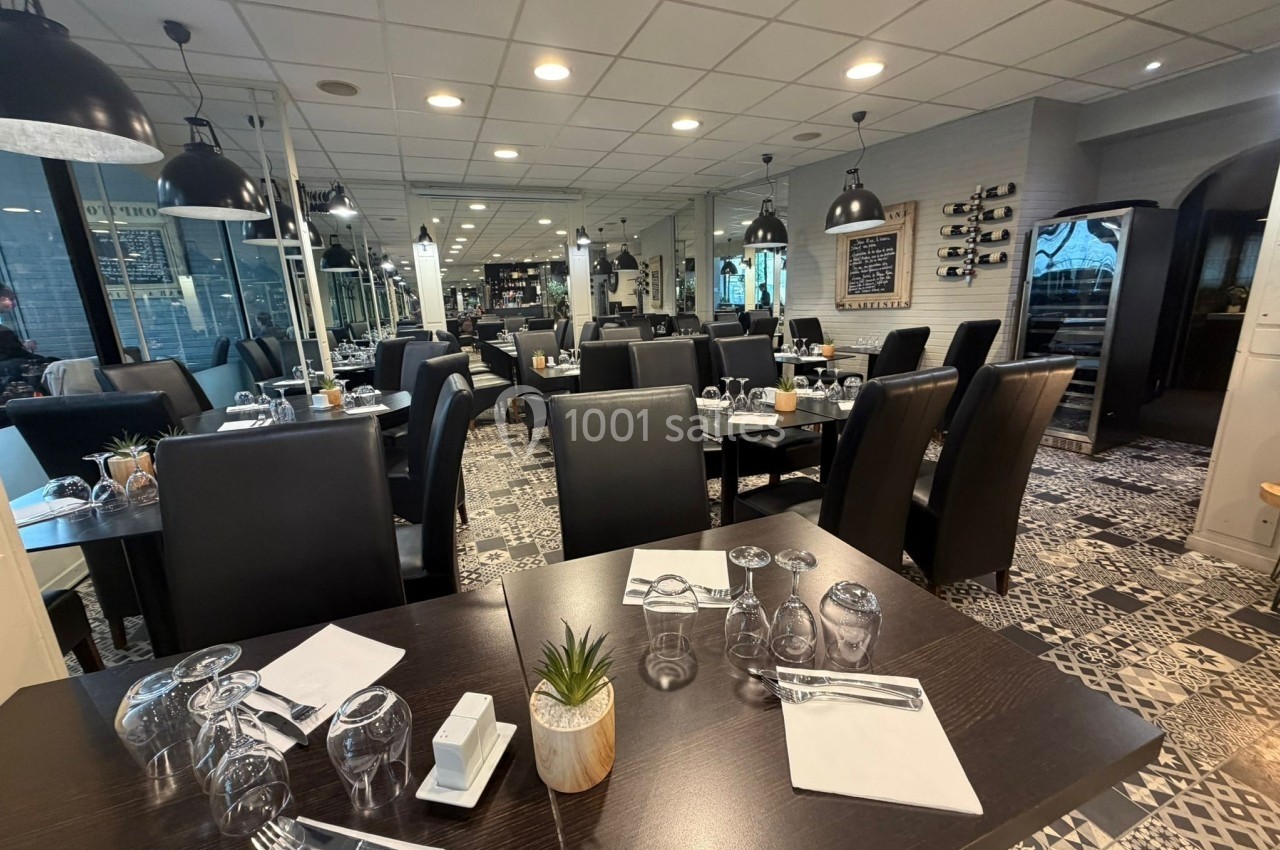 Salle de restaurant moderne avec tables dressées, chaises noires et éclairage suspendu dans un décor lumineux.
