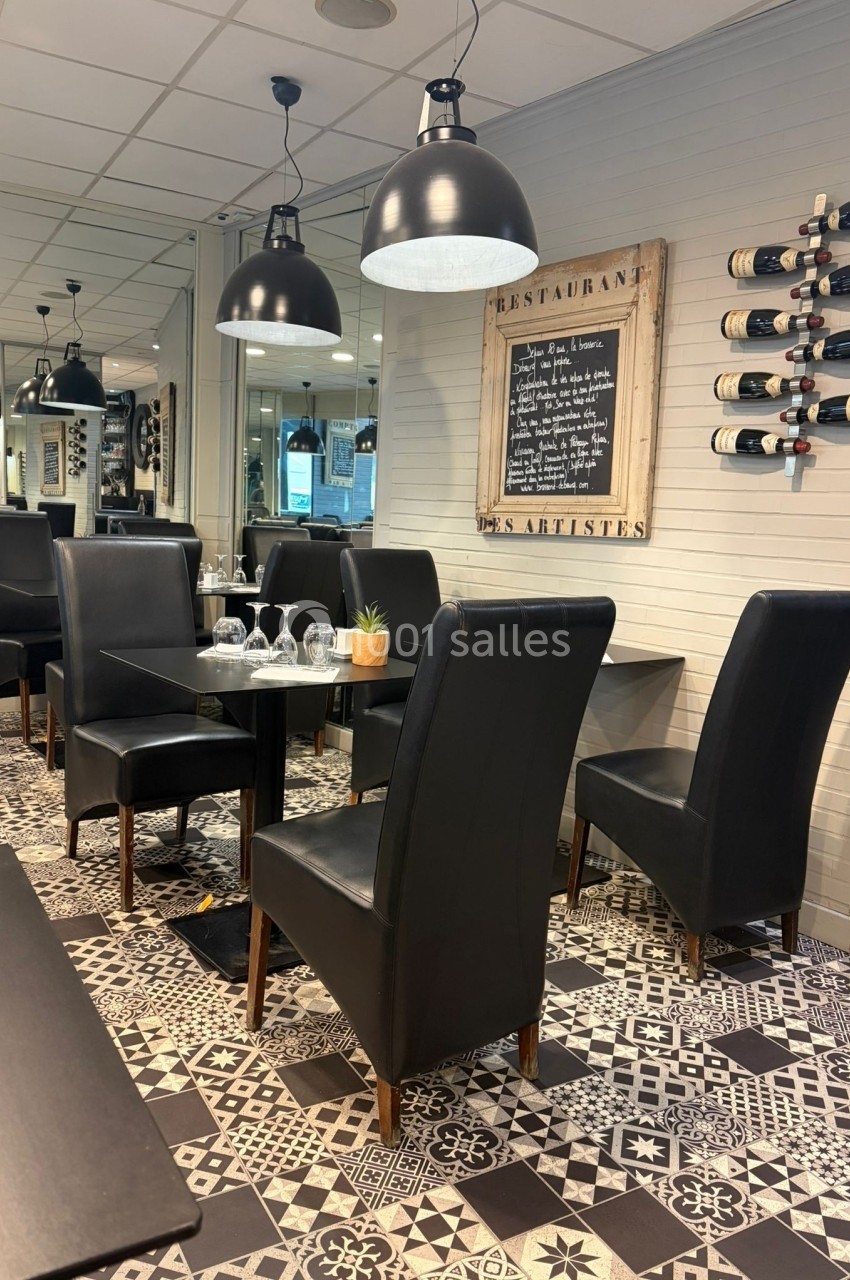 Salle de restaurant avec tables dressées, chaises noires, luminaires suspendus et décoration murale sobre.