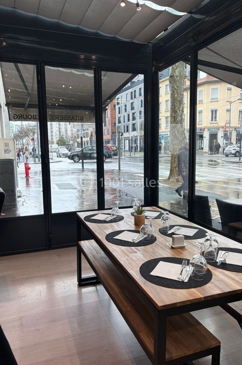 Salle de restaurant avec une table dressée près d'une baie vitrée donnant sur une rue urbaine sous la pluie.