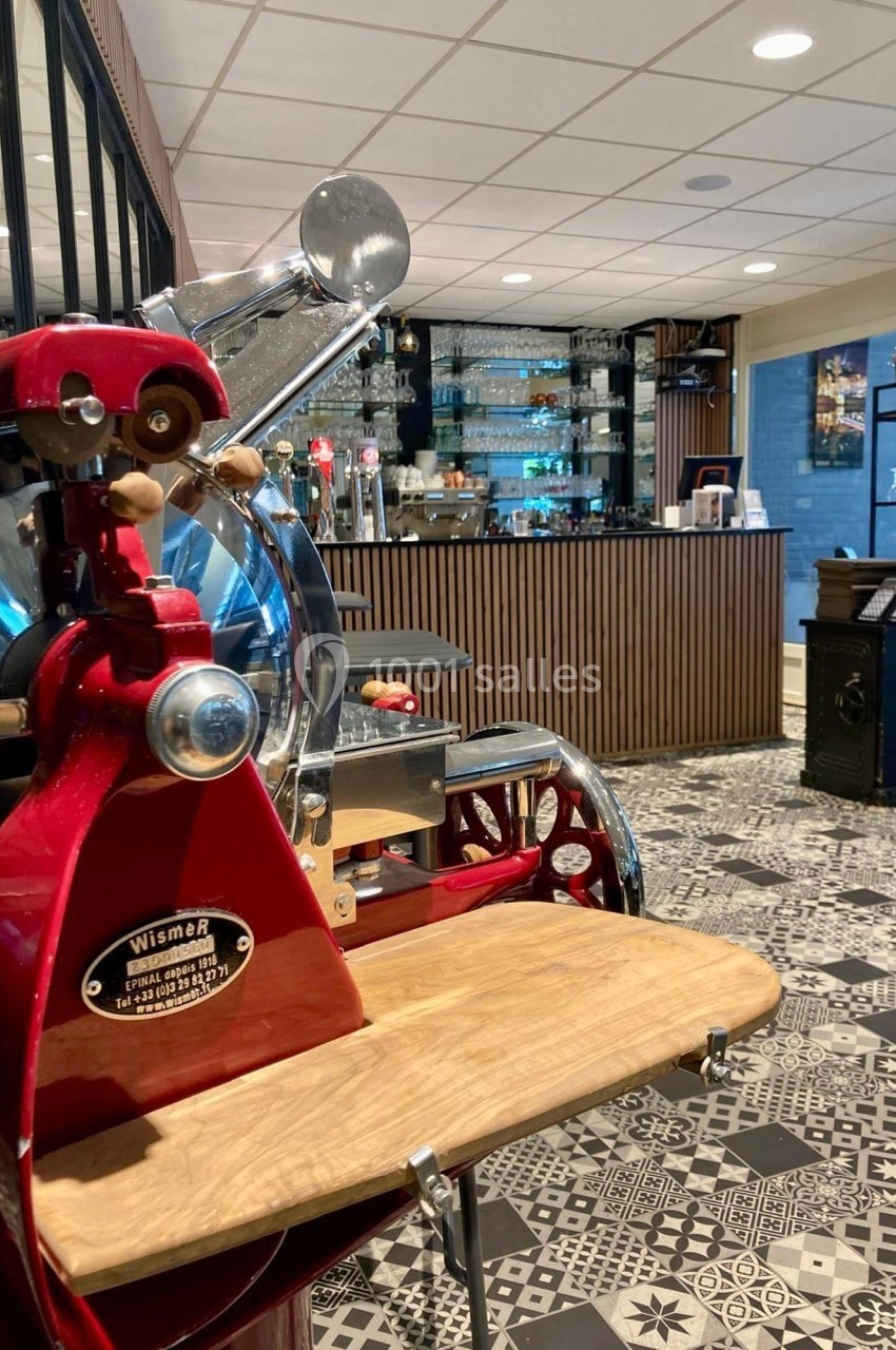 Trancheuse rouge vintage en premier plan dans un espace intérieur avec bar et sol en carreaux décoratifs.