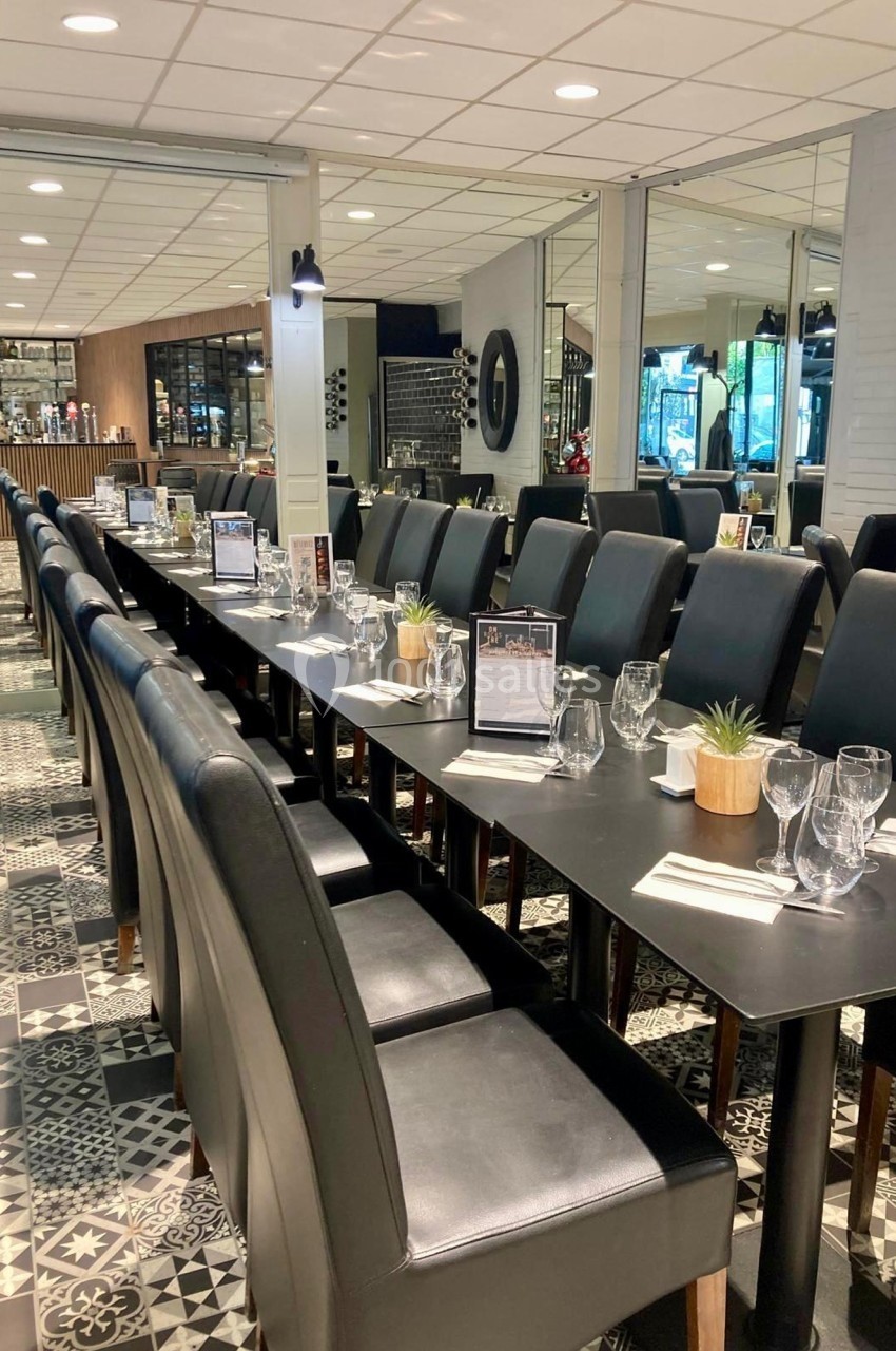 Salle de restaurant avec une longue table dressée, chaises en cuir noir et décoration moderne.