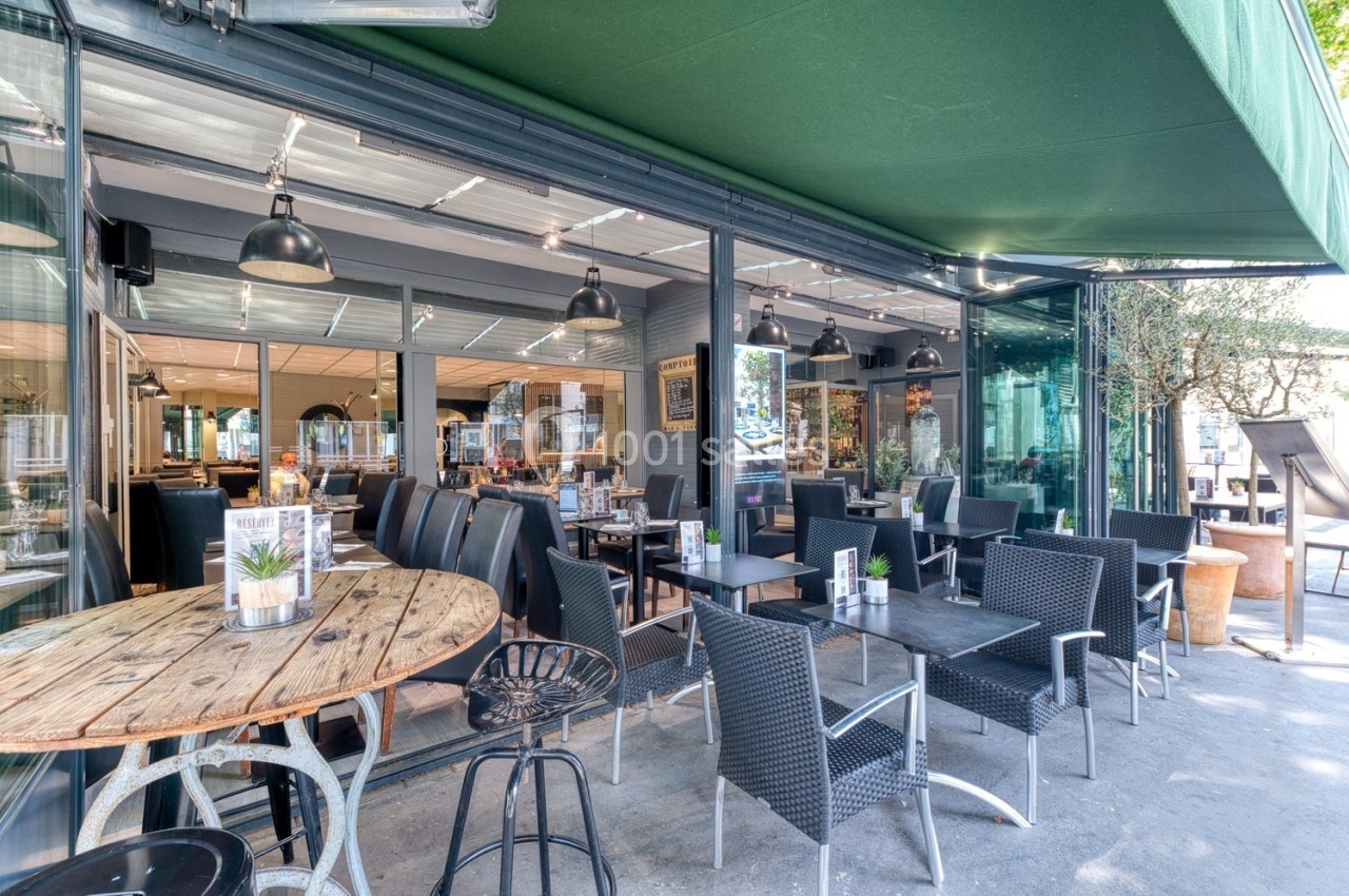 Terrasse d'un café-restaurant avec tables en bois et en rotin, sous un auvent vert, donnant sur l'intérieur vitré.