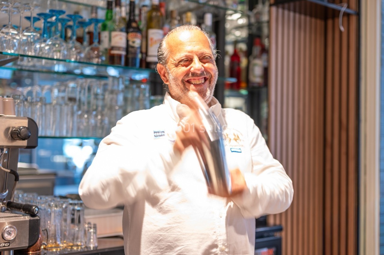 Un barman souriant agite un shaker derrière un comptoir avec des étagères remplies de bouteilles et de verres.