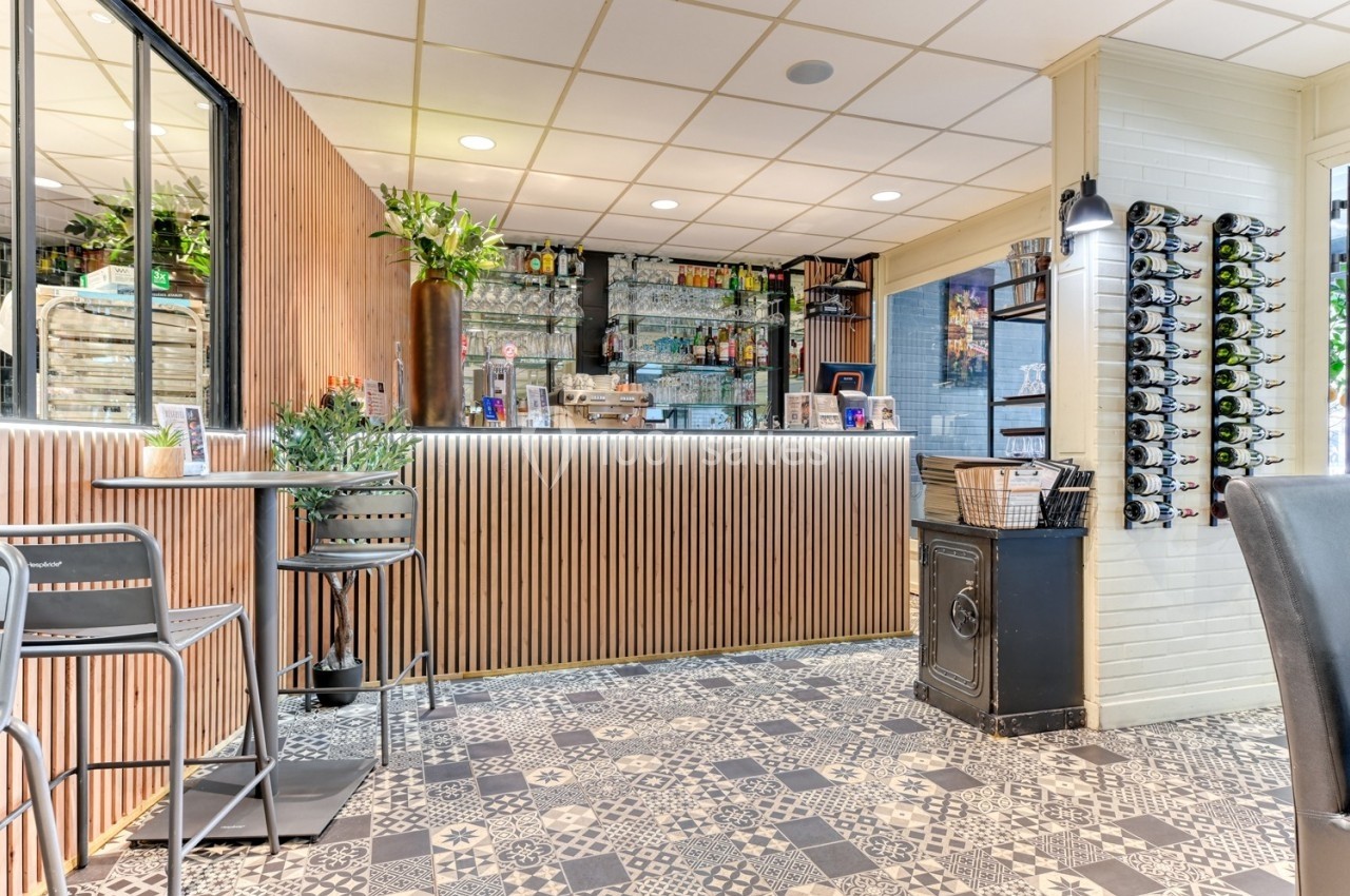 Comptoir de bar moderne avec revêtement en bois, étagères de bouteilles, tables hautes et décoration végétale.