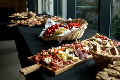 Plateaux de charcuterie, fromages et pains disposés sur une table avec des paniers de fruits et légumes en arrière-plan.