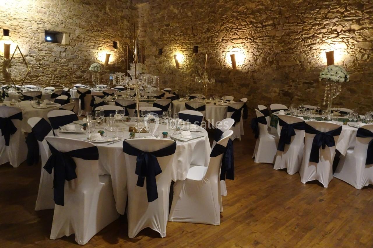 Salle de réception avec murs en pierre, tables rondes dressées et chaises blanches décorées de nœuds noirs.