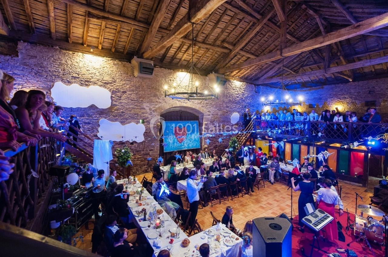 Salle en pierre avec plafond en poutres, tables dressées pour un repas, invités assis et musiciens sur scène.