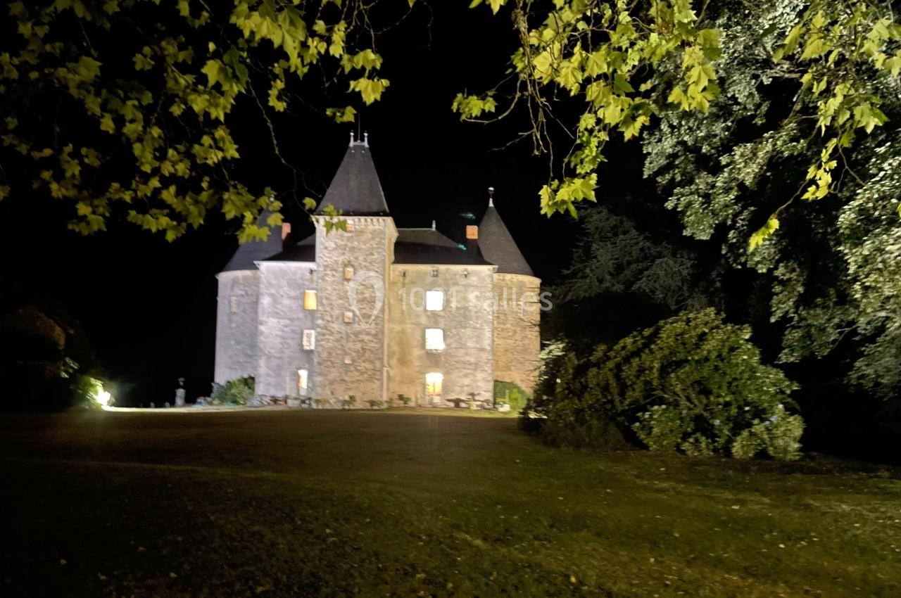 Château en pierre éclairé de nuit, entouré d'arbres et d'une pelouse sombre au premier plan.