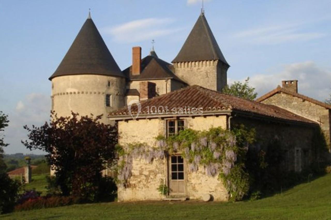 Château médiéval avec tours rondes, petite maison en pierre couverte de glycine, entourés de verdure sous un ciel dégagé.