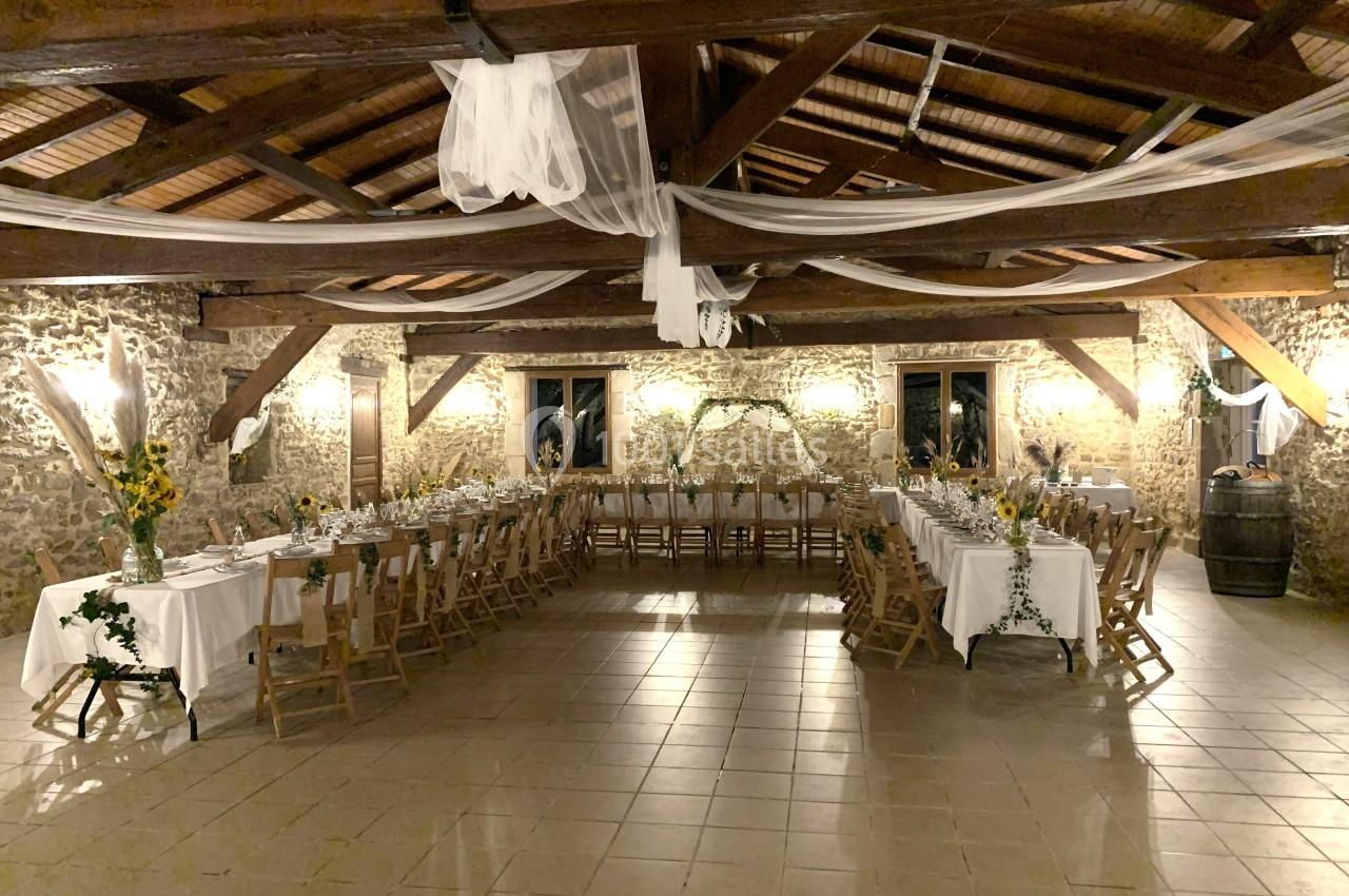 Salle de réception rustique avec tables décorées de nappes blanches, tournesols et guirlandes, sous un plafond en poutres…