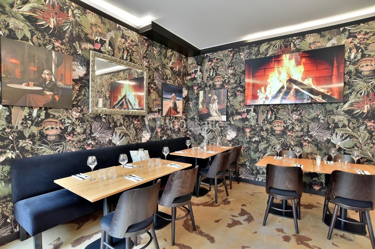 Salle de restaurant avec tables en bois, banquettes noires, décoration murale tropicale et écran affichant une cheminée.