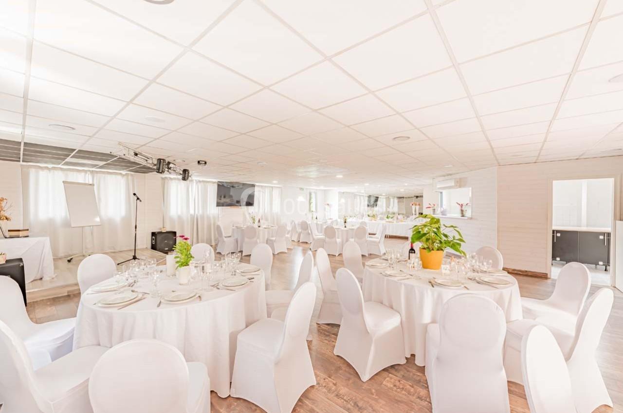 Salle de réception lumineuse avec tables rondes dressées, nappes blanches et chaises couvertes, décorée de plantes.