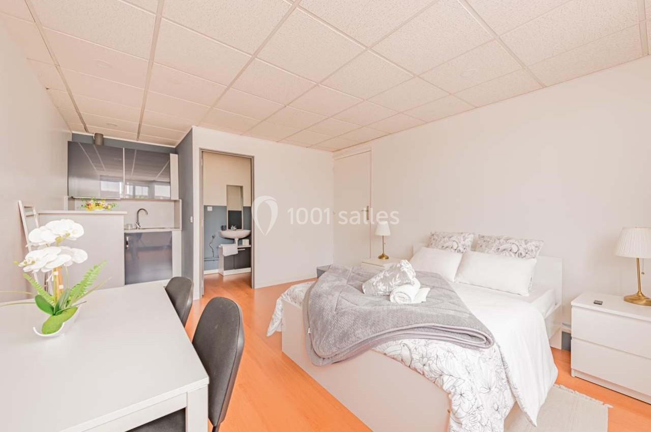 Chambre lumineuse avec lit double, bureau, kitchenette et salle de bain attenante dans un espace moderne et épuré.