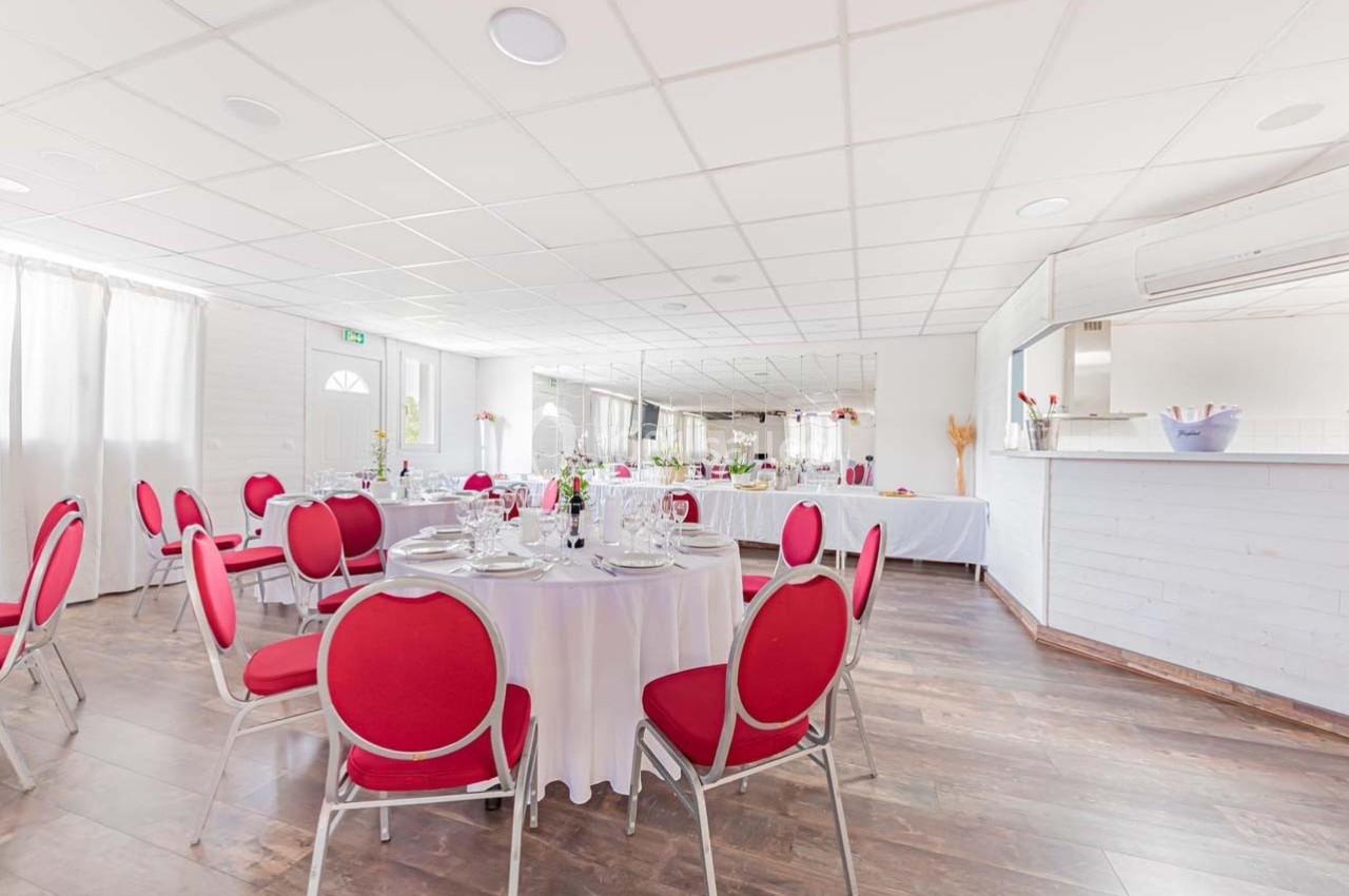 Salle lumineuse avec tables rondes dressées, chaises rouges, et buffet en arrière-plan près d'un comptoir blanc.