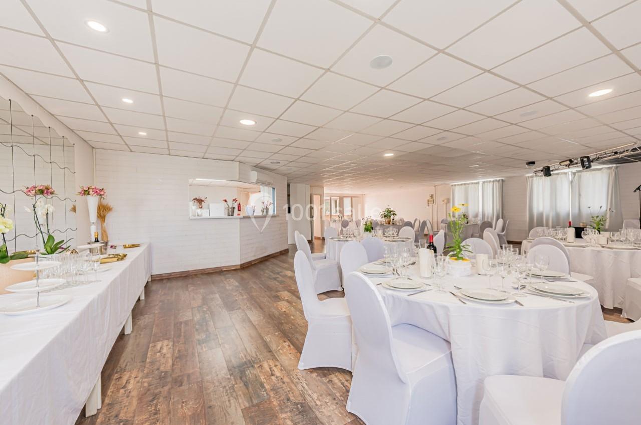 Salle de réception lumineuse avec tables rondes dressées, nappes blanches et décorations florales.