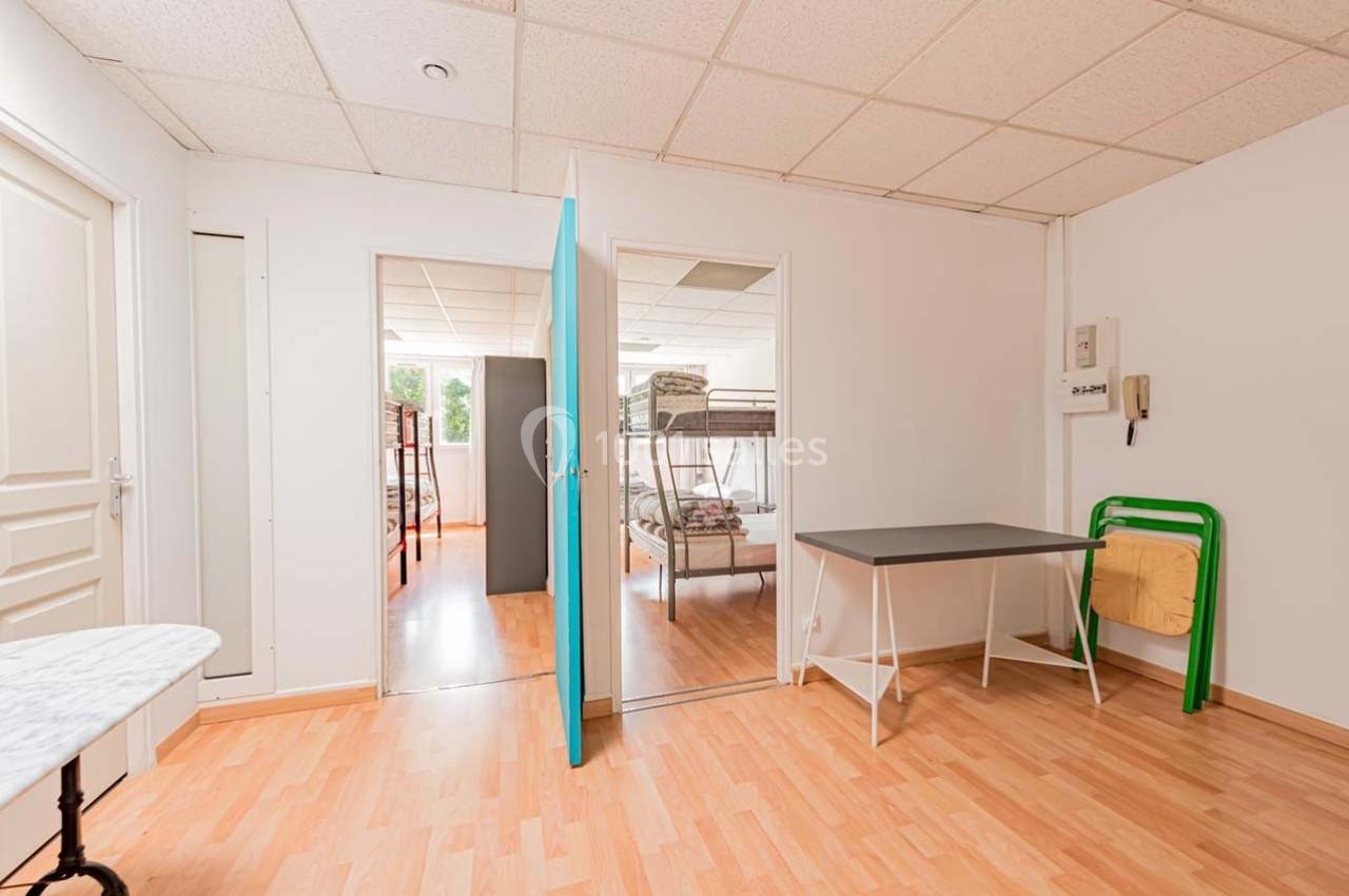 Salle lumineuse avec sol en parquet, comprenant une table, des lits superposés visibles à travers une porte ouverte.