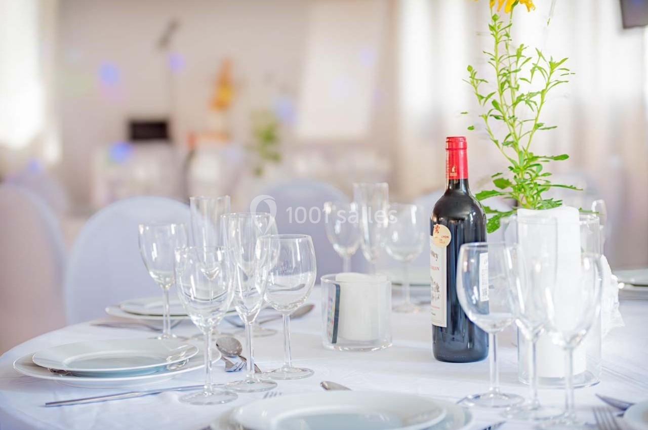 Table élégamment dressée avec nappes blanches, verres, assiettes et une bouteille de vin rouge au centre.