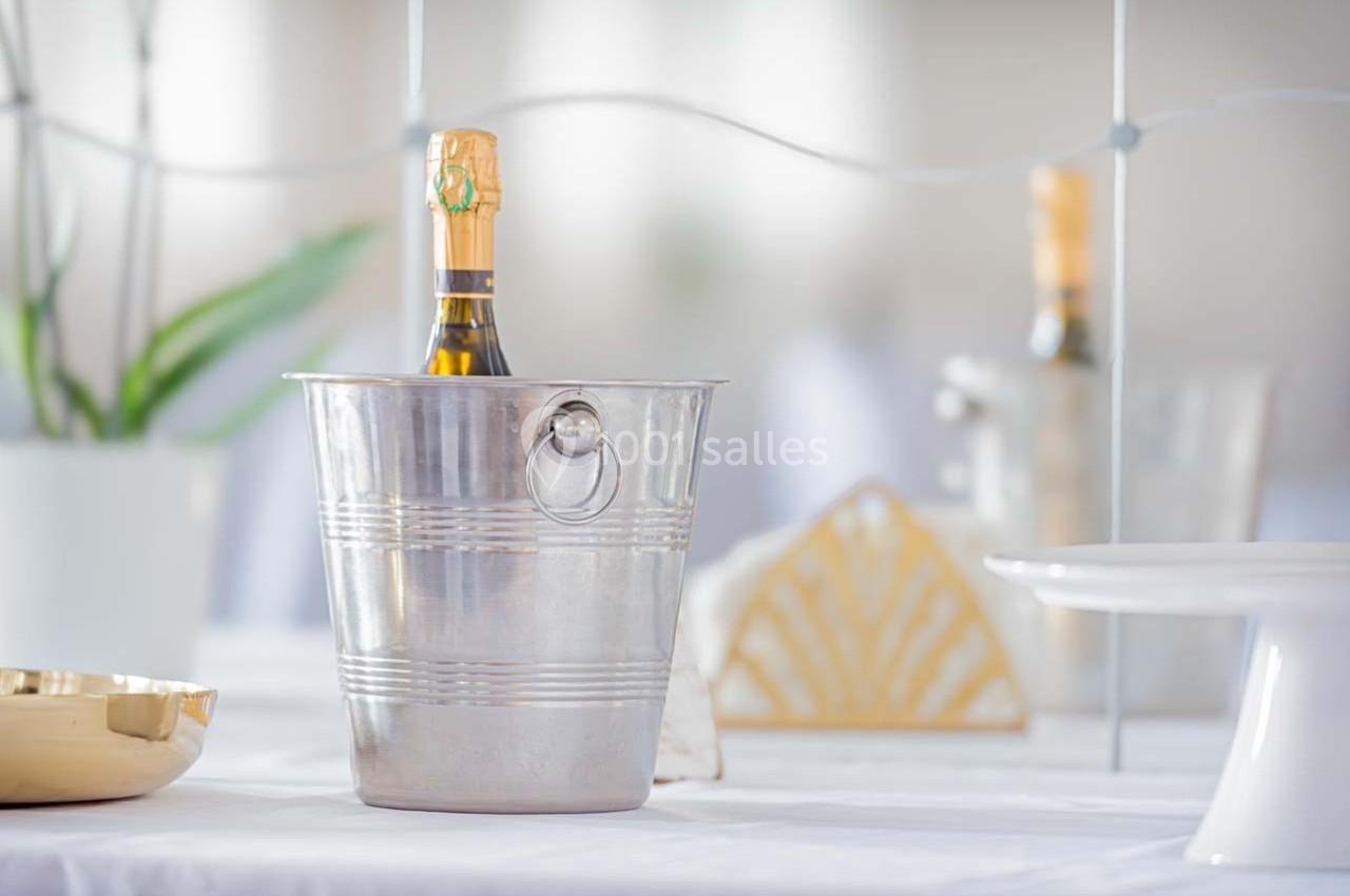 Bouteille de champagne dans un seau en métal posé sur une table blanche, avec des éléments de décoration en arrière-plan.
