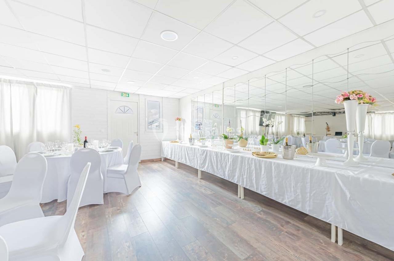 Salle lumineuse avec tables rondes dressées et buffet décoré, sol en bois et murs ornés de miroirs.