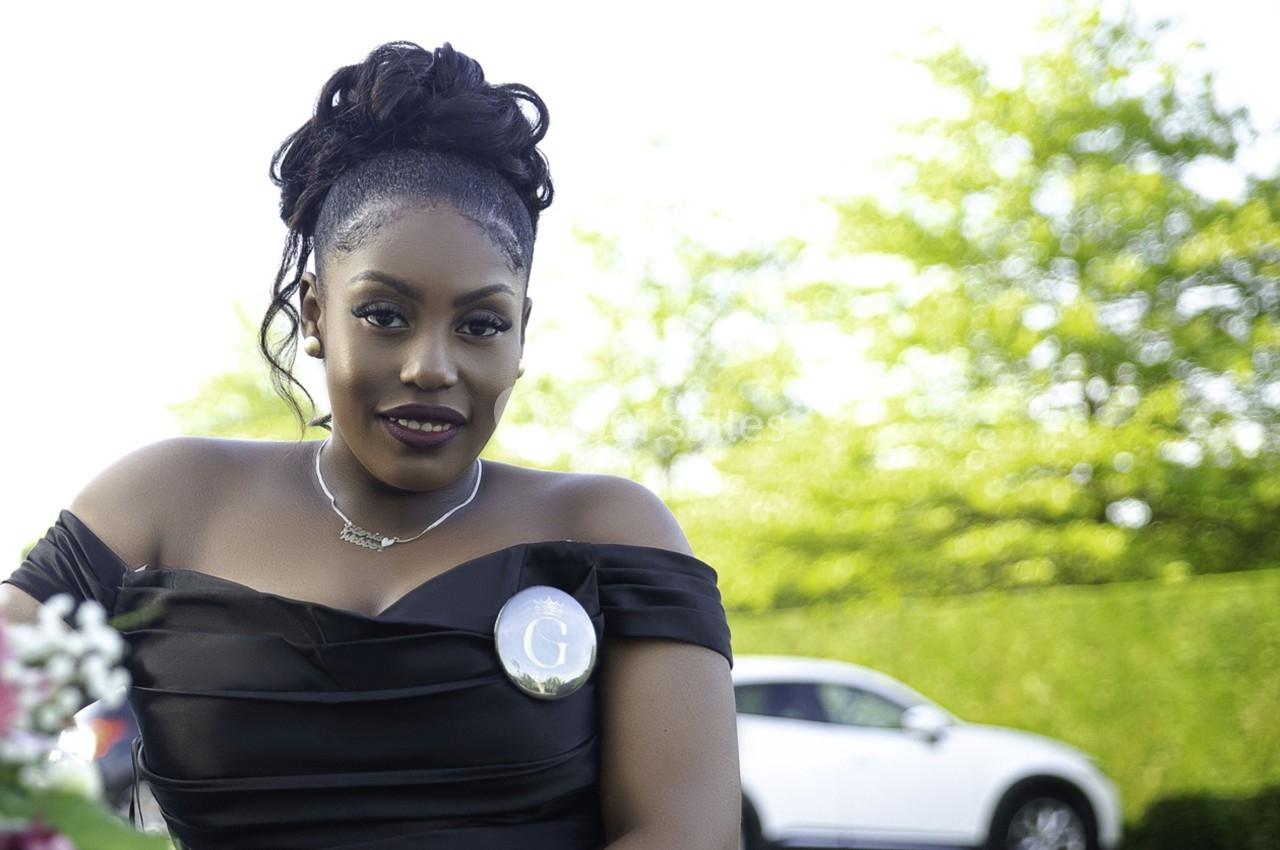 Femme souriante en robe noire, portant un badge, assise en extérieur avec une voiture et des arbres en arrière-plan.