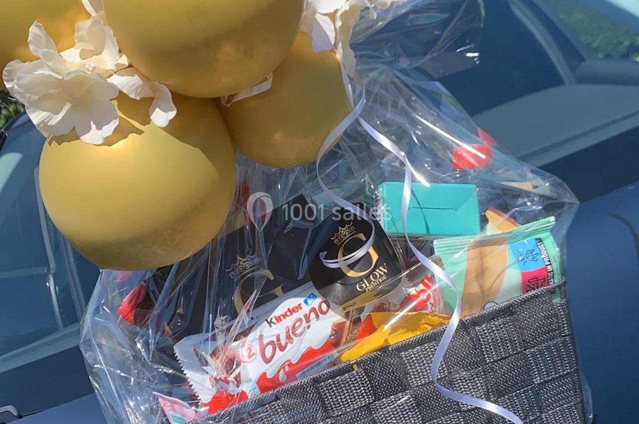 Panier cadeau contenant des friandises, des ballons dorés et des décorations, posé sur une voiture.