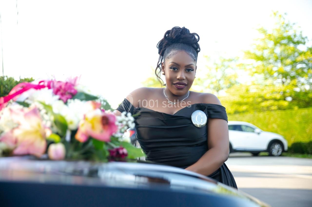 Femme en robe noire posant à l'extérieur près d'un bouquet de fleurs, avec des voitures en arrière-plan.