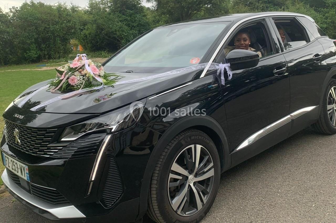 Voiture noire décorée de fleurs et de rubans blancs, utilisée pour une cérémonie, avec des passagers à l'intérieur.