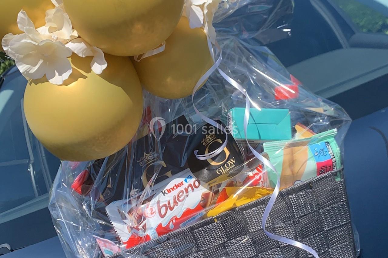 Panier cadeau contenant des friandises, des produits divers et des ballons dorés, posé sur une voiture.