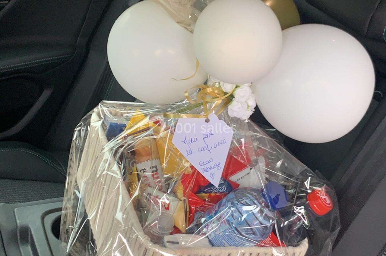 Panier cadeau emballé avec des produits variés, décoré de ballons blancs et dorés, posé sur un siège de voiture.