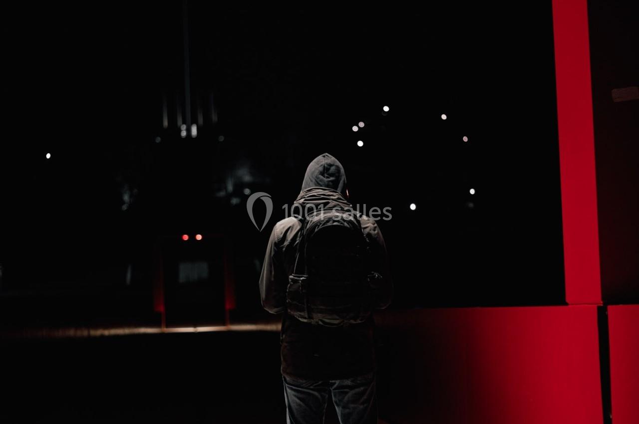 Un individu en capuche et sac à dos se tient seul dans un environnement urbain sombre, éclairé par des lumières rouges.