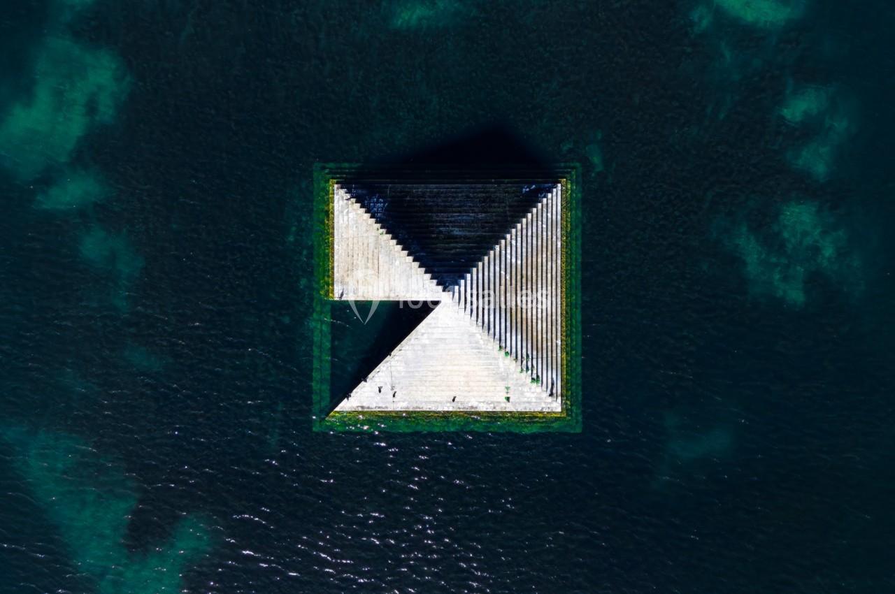 Vue aérienne d'une structure géométrique flottante en forme de triangle et carré sur une étendue d'eau sombre.