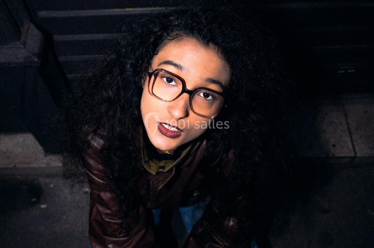 Jeune femme aux cheveux bouclés et lunettes, accroupie sur un trottoir devant un mur sombre avec des graffitis jaunes.
