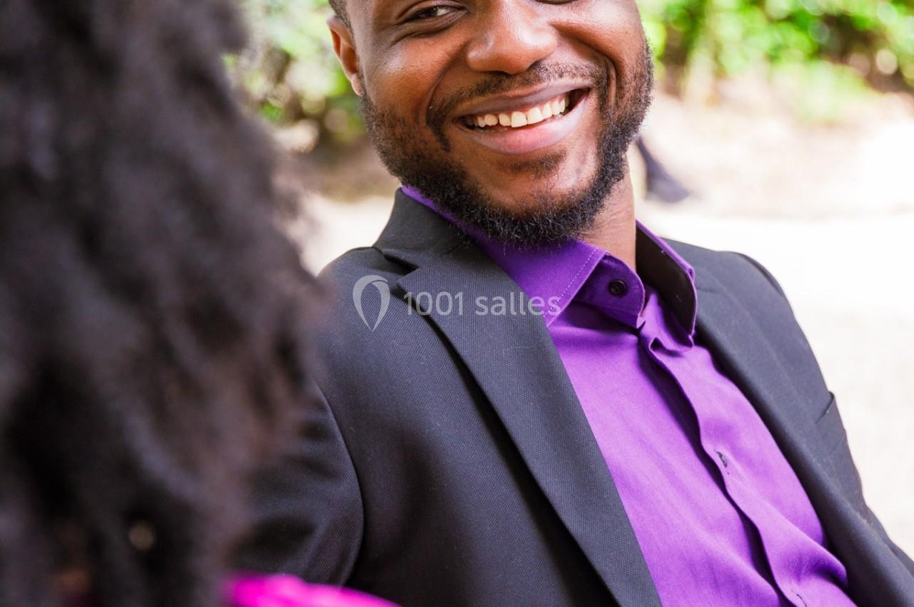 Homme souriant en costume noir et chemise violette, assis en extérieur, discutant avec une personne de dos.