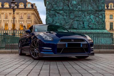 Voiture sportive bleue stationnée sur une place pavée devant un monument en bronze et des bâtiments historiques.