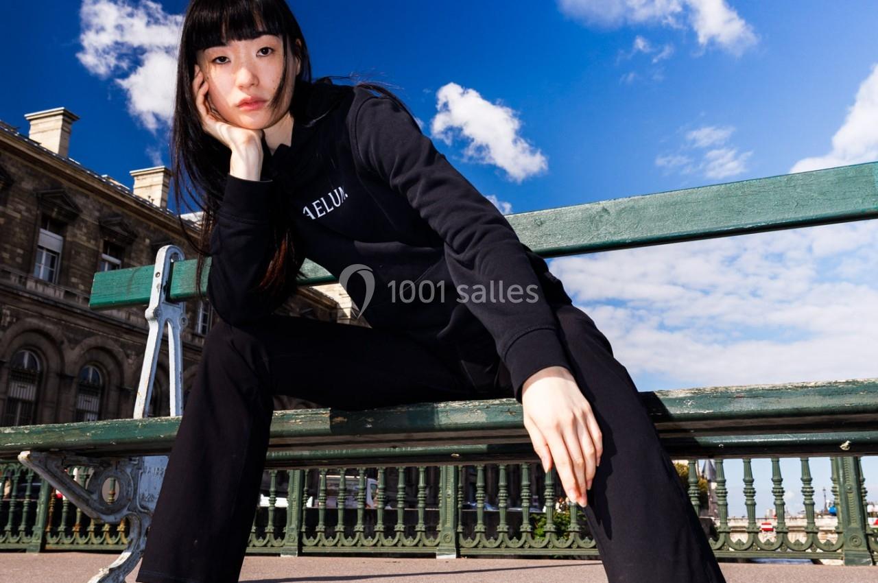 Femme assise sur un banc en extérieur, portant des vêtements noirs, ciel bleu et bâtiment en arrière-plan.