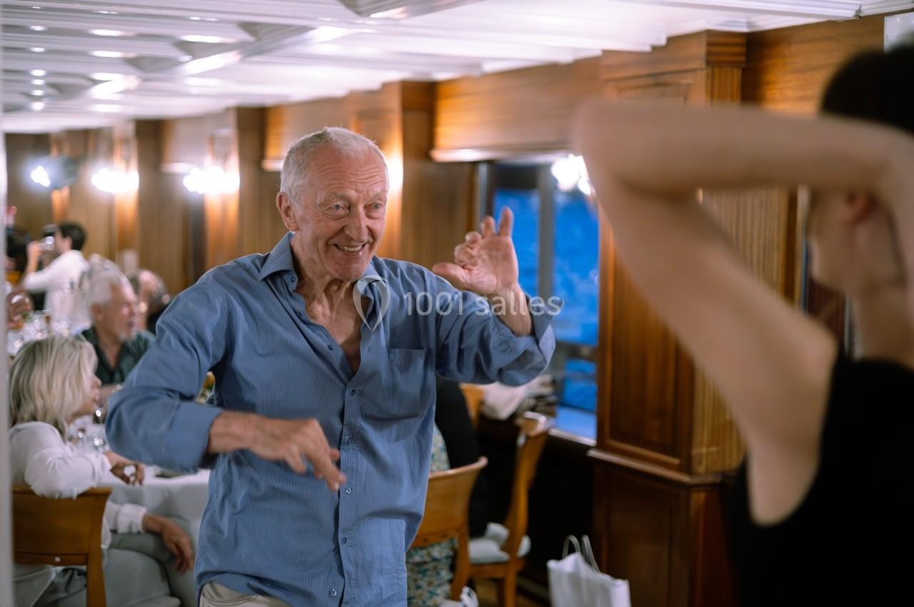 Un homme âgé danse joyeusement dans une salle en bois éclairée, entouré de personnes assises.